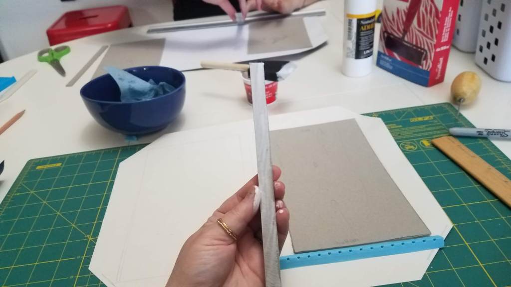Photo of a workspace that includes a teal cutting mat which has cut paper and a rectangle of cardstock on top, a wooden 12-inch ruler, a black Sharpie marker, an awl, a white plastic storage container, a box containing a brayer, a bottle of fancy white glue, a yogurt cup containing glue and a a foam paintbrush covered in glue at the tip, a blue bowl with a wet, blue paper towel wadded up inside of it, a pair of green-handled scissors, a red stool, another work area across the table from the main work area (but without the teal mat), and front-and-center a hand which is coated in glue holding a cardstock spine that is also coated in glue. So much glue.