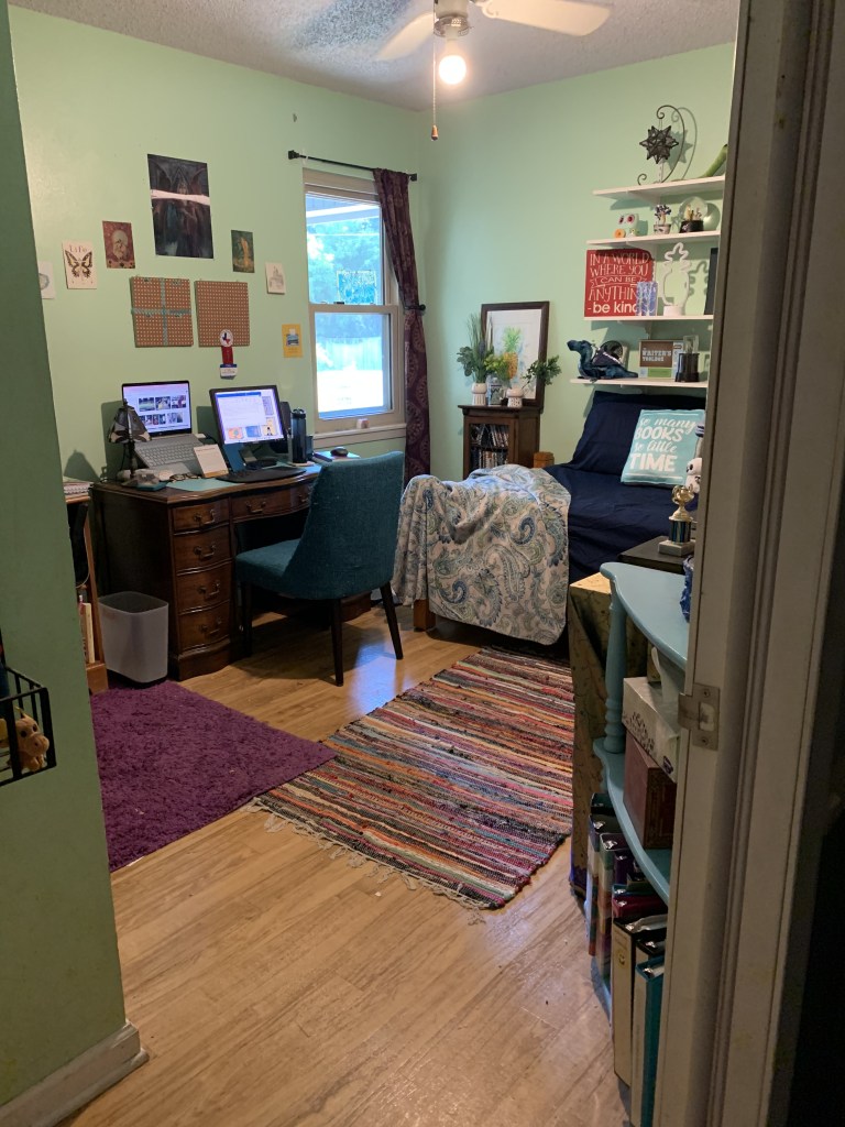 Looking into a small home office space from a doorway. There is a window in the center of the frame. to the left is a dark wooden desk with nine drawers. It has a laptop with an extra monitor on top, plus a tiffany style lamp on the left side. In front of the desk is a teal fabric chair. On the wall above the desk is a couple of small square corkboards and some small card-sized pieces of art with fairies and dragons. To the right of the window is a three foot tall, narrow bookshelf filled with small spiral notebooks. Sitting on top of it is two small white vases filles with greenery and a large watercolor painting of a pineapple. To the left of the shelf is a twin size bed draped in a white blanket with blue, green, and teal paisley. The sheet and pillows on the bed are navy. Above the bed are four white, floating shelves, which have dragons, pineapples, a writer's block emergency kit, and a many pointed star lamp. 