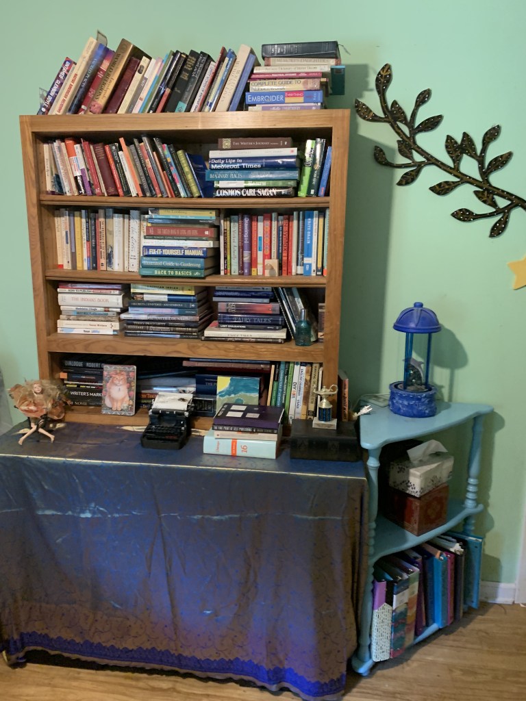 In front of a mint green wall, a three foot tall by four foot wide golden wood bookcase is sitting on top of something draped in gold and blue fabric. The bookshelf is filled every which way with books. A small orange fairy, a small black typewriter, and a stack of books sit on the fabric. To the right of the bookcase, on the wall, a decorative black branch with many leaves and gold paint hangs above a yellow ceramic star-shaped light switch cover. Below that sits a three-tier corner shelf that has been painted turquoise. On the top shelf is a dolphin oil lamp. The second shelf contains a red square box and a box of tissues. The bottom shelf holds many different colored binders.